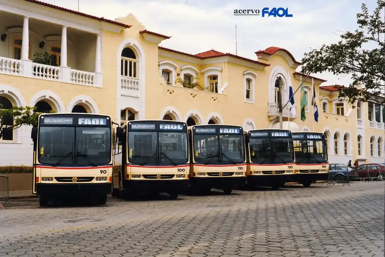 Cinco ônibus da FAOL alinhados diante de um prédio amarelo com janelas e arcos brancos.