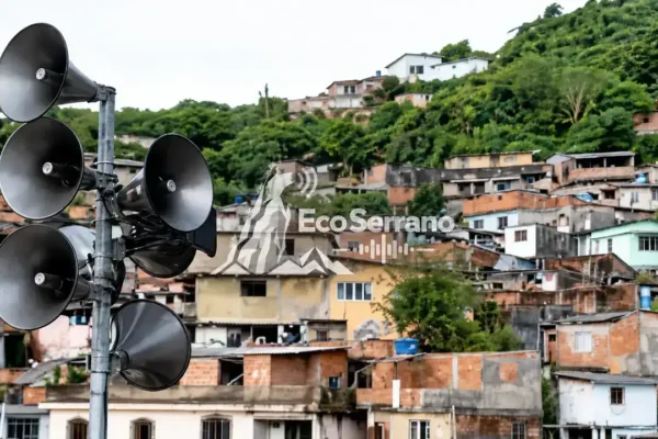 Sirenes instaladas em poste, com casas e vegetação em uma encosta ao fundo.