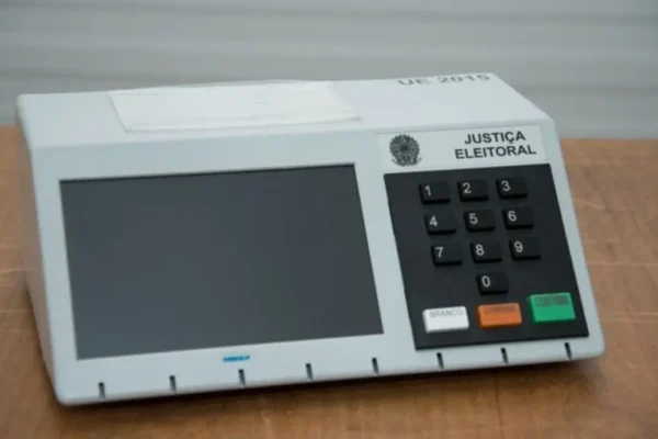 Urna eletrônica sobre mesa de madeira, com tela escura e teclado numérico à direita.