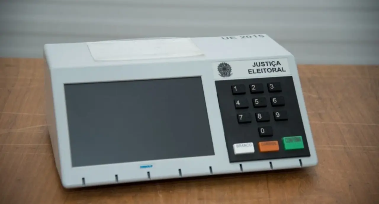 Urna eletrônica sobre mesa de madeira, com tela escura e teclado numérico à direita.