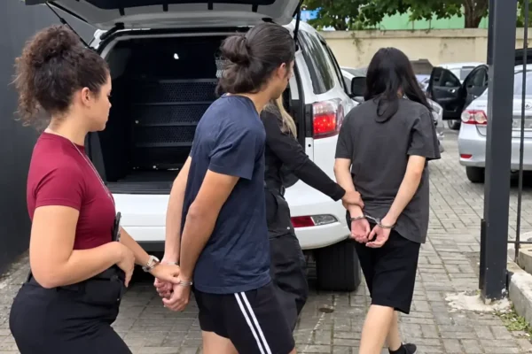 Duas pessoas algemadas são conduzidas por agentes ao lado de um veículo branco.