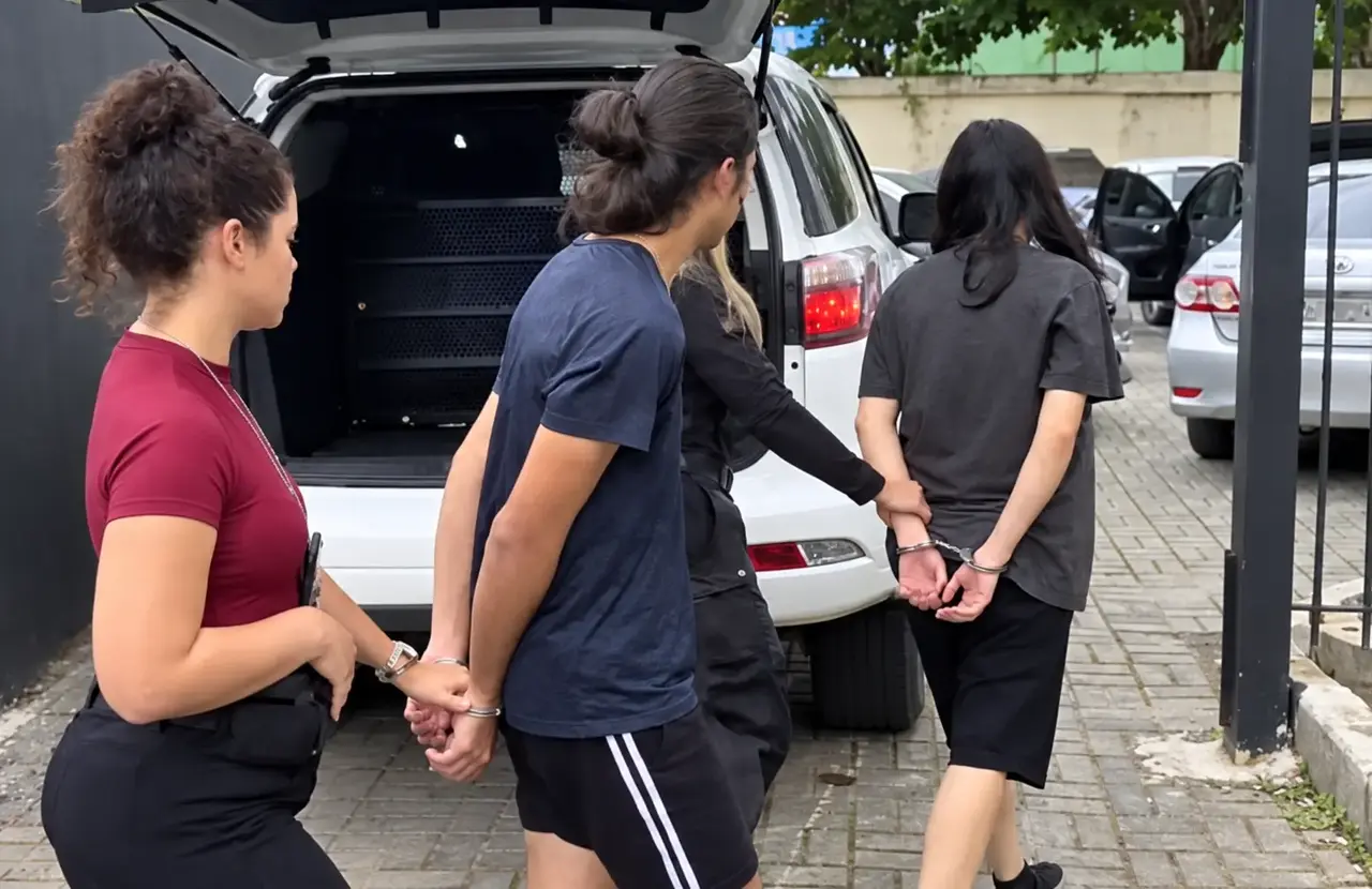 Duas pessoas algemadas são conduzidas por agentes ao lado de um veículo branco.