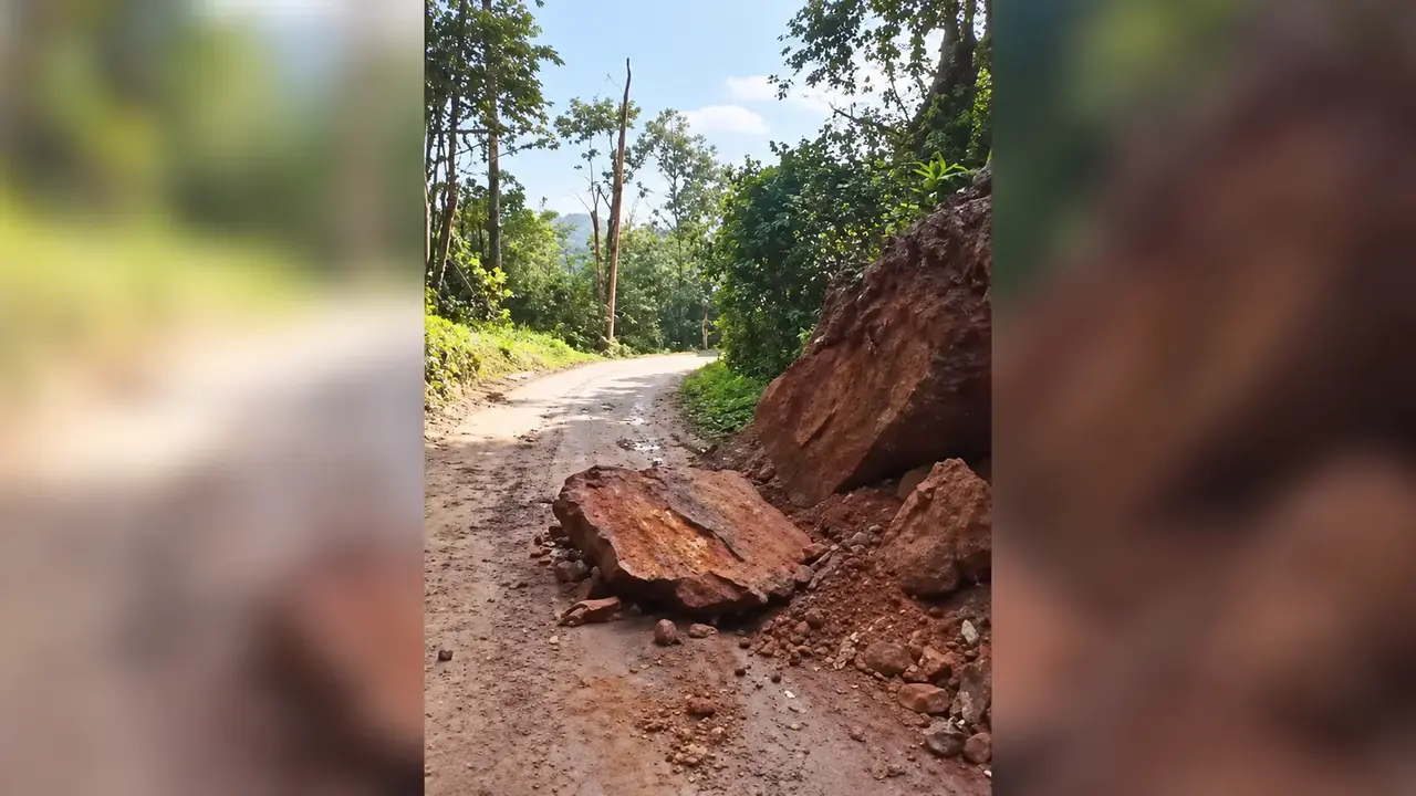 Pedra de grande porte e terra sobre parte de uma estrada rural, junto a uma encosta cercada por vegetação.