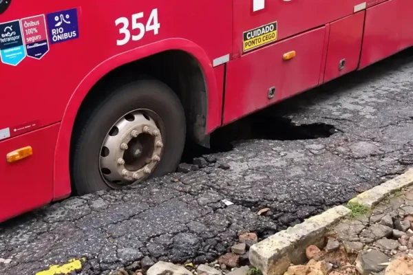 Ônibus vermelho com uma roda afundada em buraco no asfalto, próximo ao meio-fio.