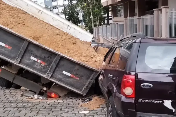 Caminhão carregado de terra inclinado em um buraco na rua, junto a um carro escuro.