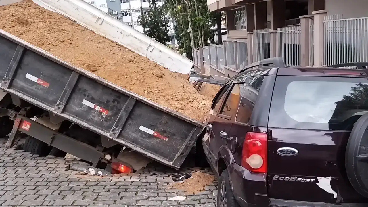 Caminhão carregado de terra inclinado em um buraco na rua, junto a um carro escuro.